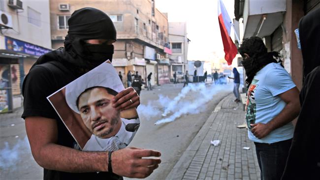 This file photo shows a masked Bahraini anti-government protester holding a picture of jailed Shia cleric Sheik Ali Salman as riot police fire tear gas canisters during clashes in Bilad Al Qadeem, Bahrain. (By AP)
