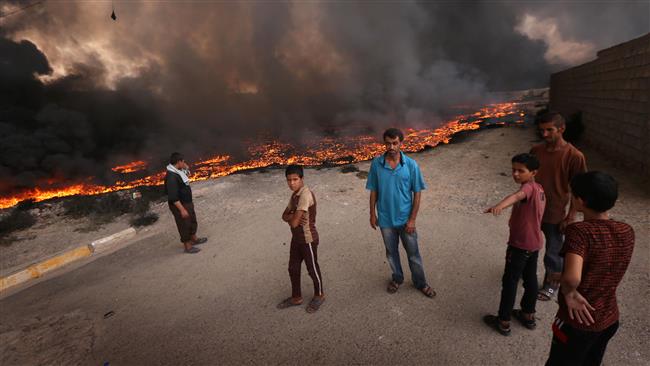 People stand near fire and smoke billowing from oil wells set ablaze by Daesh Takfiri terrorists before fleeing the oil-producing region of Qayyarah, on August 30, 2016, after Iraqi forces pushed the militants out of the northern town on the banks of the Tigris river